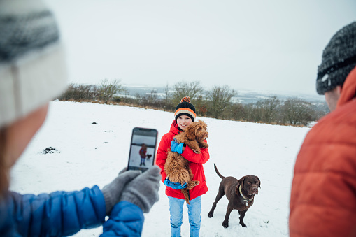 An unrecognisable caucasian woman taking a photo of her son and two dogs on her smart phone. They are wearing warm winter clothing and accessories in a snowy, rural, outdoor setting.