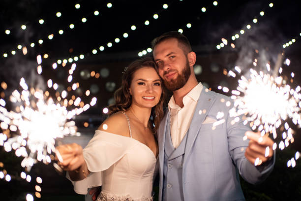 newlyweds with sparklers in night park with garlands in trees. ending to wedding newlyweds with sparklers in a night park with garlands on the trees. A perfect end to the wedding day. shooting in the dark. the bride in a white elegant dress and the groom in a blue suit. wedding fireworks stock pictures, royalty-free photos & images