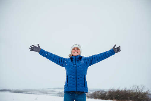 A mature caucasian woman wearing warm winter clothing and accessories in a snowy, rural, outdoor setting. She is standing in the snow with her arms outstretched.