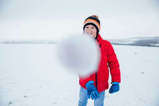 A caucasian boy wearing warm winter clothing and accessories in a snowy, rural, outdoor setting. He is throwing a snowball at the camera.