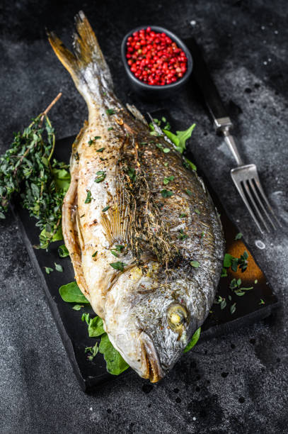 istock Roasted sea bream  fish with herbs on a cutting board. Black background. Top view 1303692688