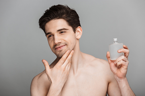 Beauty portrait of half naked handsome young man using aftershave lotion isolated over gray background