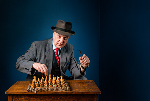 A middle aged senior man in suit sits alone at a wooden table, playing a game of chess