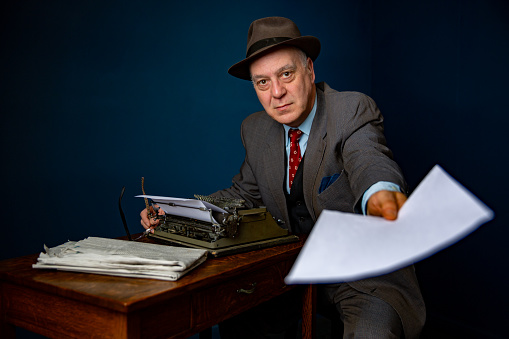 Friendly senior mature man in suit and hat sits at wooden desk with vintage typewriter and paper, handing over an empty page