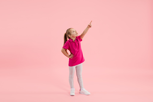 Pointing. Childhood and dream about big and famous future. Pretty little girl isolated on coral pink studio background. Dreams, imagination, education, facial expression, emotions concept.