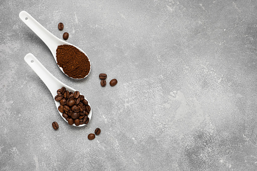 White porcelain spoons with coffee beans and ground coffee on a gray table. Top view, copy space.