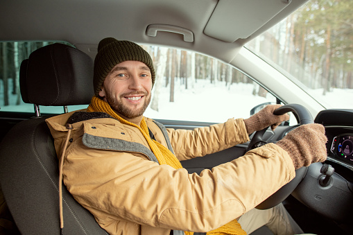 Happy young active man in warm winterwear looking at you with toothy smile while sitting by steer and driving to his country house