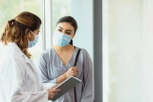 The mid adult female gynecologist explains the test results to the mid adult female patient.  The results are displayed on the digital tablet.  Both women wear masks because of COVID-19.