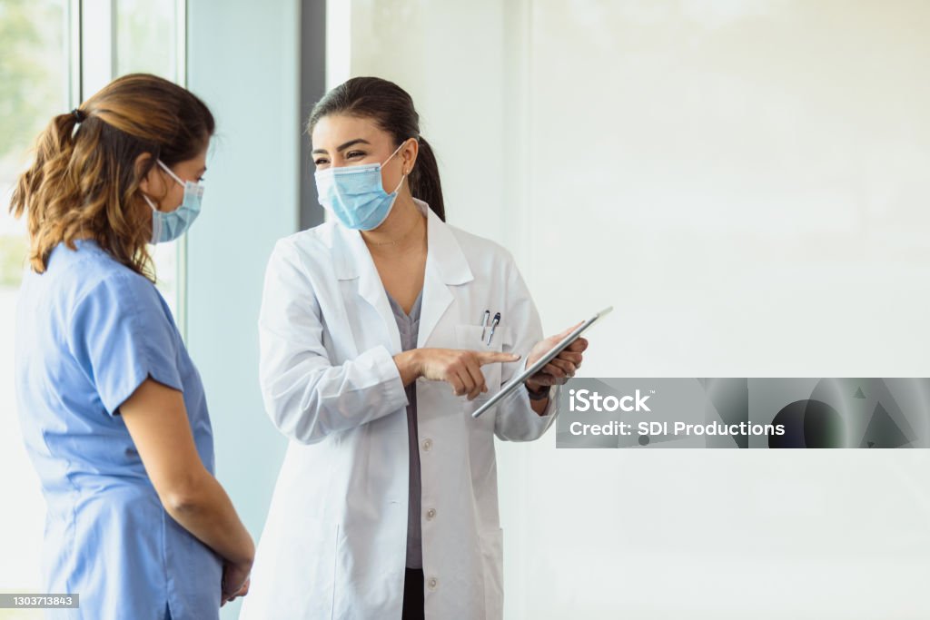 Smiling female doctor points to patient's test results on tablet The smiling mid adult female doctor shows the patient's good test results shown on the digital tablet to her mid adult female colleague.  They both wear masks to flatten the curve. Protective Face Mask Stock Photo