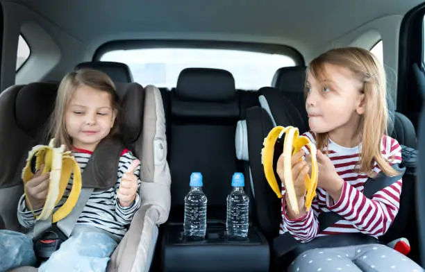 Photo of Little girls, sisters are driving in car, eating banana. Traveling on road in safe baby seats with child belts. Fun family trip, activity with parents