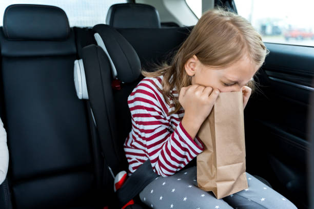 Little girl is driving in car. Kid is sick, feels bad and vomiting into paper bag. Traveling, riding on road in safe baby seats with child belts. Fun family trip, activity with parents. la niña está conduciendo en coche. el niño está enfermo, se siente mal y vomita en una bolsa de papel. viajando, cabalgando por carretera en asientos de bebé seguros con cinturones para niños. divertido viaje familiar, actividad con los padres