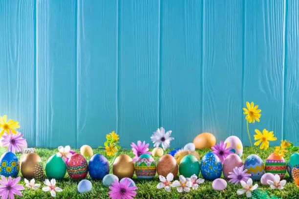 Photo of Easter eggs on turf grass and blue wooden wall with spring flowers