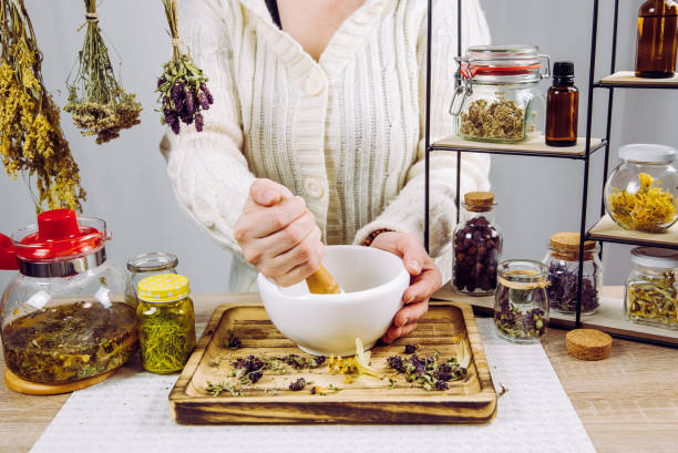 vista ravvicinata della donna erboristeria che mescola varie erbe essiccate per il tè medicinale tradizionale con mortaio e pestello. erbe essiccate in barattoli di vetro sullo sfondo. - alternative medicine mortar and pestle herbal medicine herb foto e immagini stock