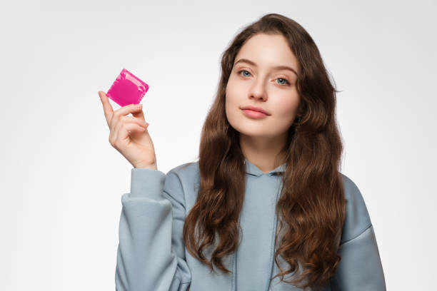 Young woman, teenager with a condom in her hand. Young woman with long hair on a light-colored background. Social advertising of condoms for young, teenagers. stock photo