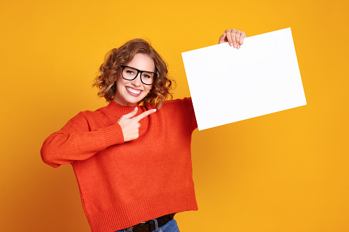 Cheerful young woman smiling for camera and pointing at empty banner against yellow background