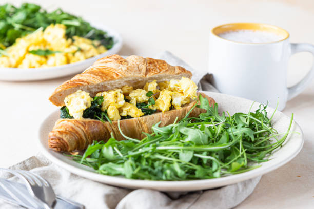 istock Delicious croissant sandwich with scrambled spinach eggs served with arugula on plate and coffee cup, light concrete background. 1303741015