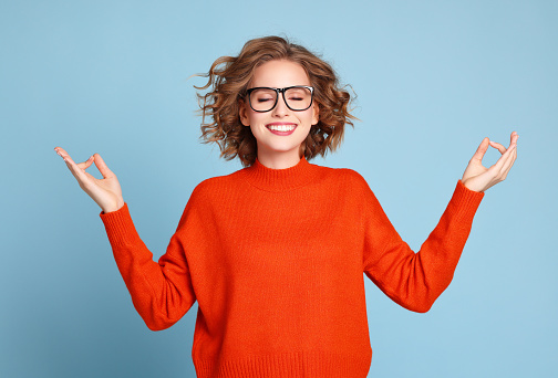 Young female in orange sweater and glasses gesturing Gyan mudra and closing eyes while meditating against blue background