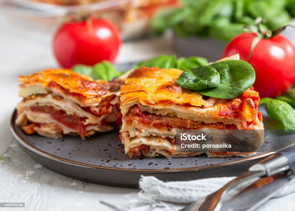Italian lasagna with tomato sauce and cheese served with tomatoes and spinach, light concrete background. Homemade vegetarian lasagna. Italian lasagna with tomato sauce and cheese served with tomatoes and spinach, light concrete background. Homemade vegetarian lasagna. Selective focus. Lasagna Stock Photo
