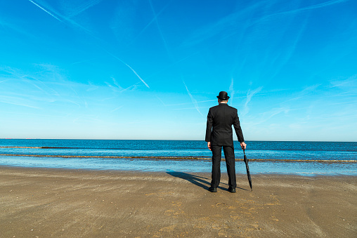 Englishman with bowler hat and umbrella stands on the beach overlooking the sea and towards Europe.