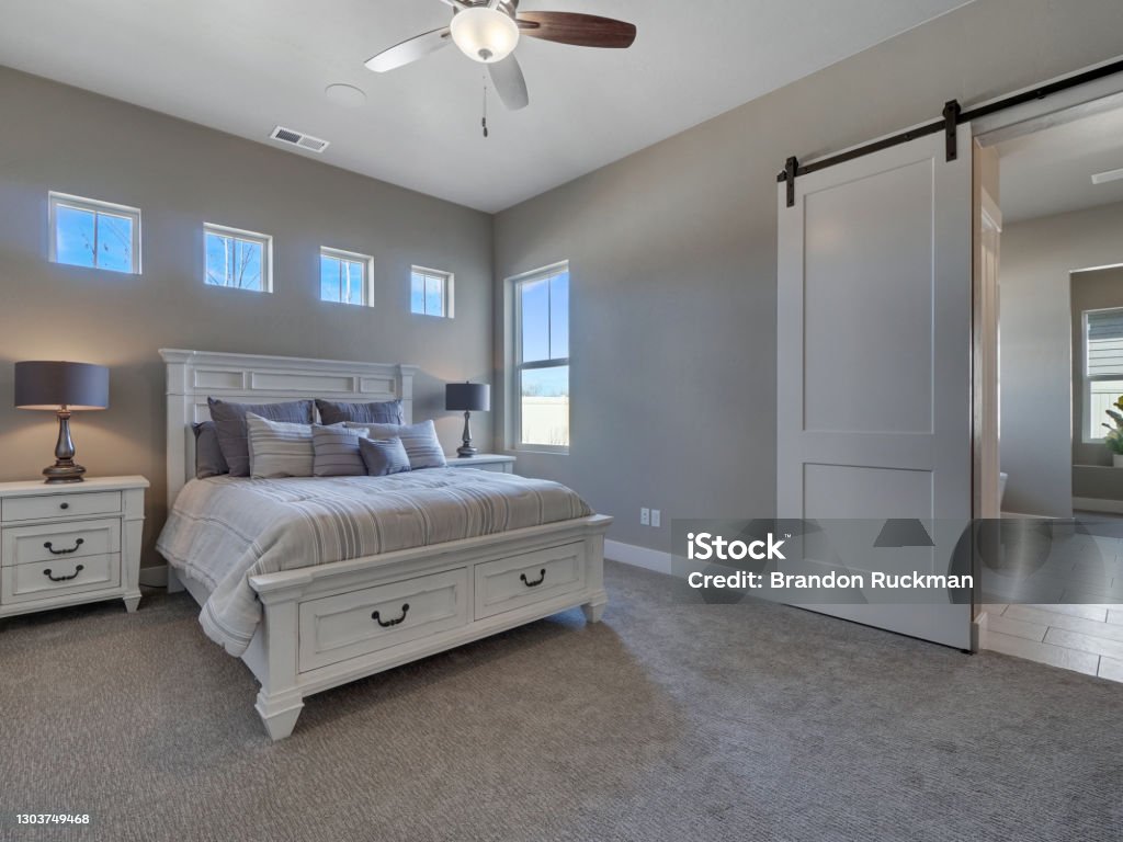 Modern Master Bedroom Staged With New White Furniture Modern Master Bedroom with new furniture and sliding barn door leading to master bathroom with windows above bed Owner's Bedroom Stock Photo