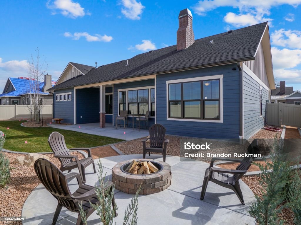Modern Backyard Firepit with Patio Furniture Real Estate Back Exterior Blue and White With landscaping modern landscaped new construction home with blue sky and puffy white clouds. Covered back patio. Features new landscaping and a fire pit with chairs surrounding it. Back Yard Stock Photo