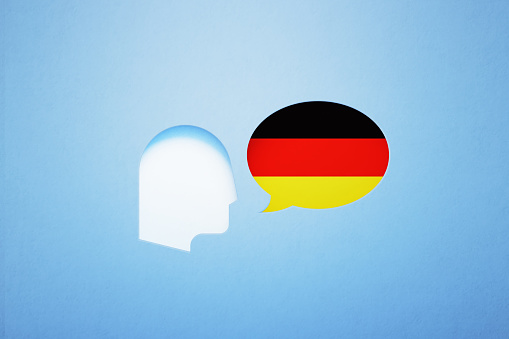 Speech bubble shape textured with German flag sitting next to a cut out human head on blue background. Horizontal composition with copy space. German learning and speaking concept.