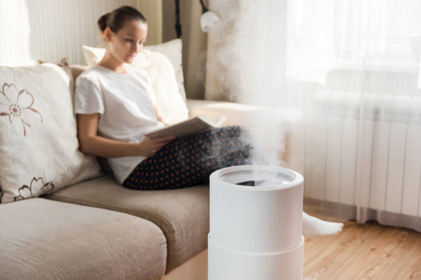 Modern air humidifier, aroma oil diffuser at home. Improving the comfort of living in a house, Improving the well-being. Ultrasonic steam technology. Woman reading book on background stock photo