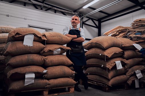 Cheerful bearded man wearing apron posing standing confidently with arms crossed near coffee bags. Full length.