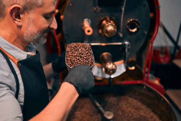 istock Side view smiling unshaved master smelling coffee grains while keeping them in ladle. He standing near industrial technical equipment. Factory concept. 1303762874