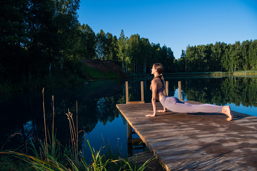 Young Woman practicing yoga on the wooden berth at lake. Single sport healthy training on nature at sunny weather.