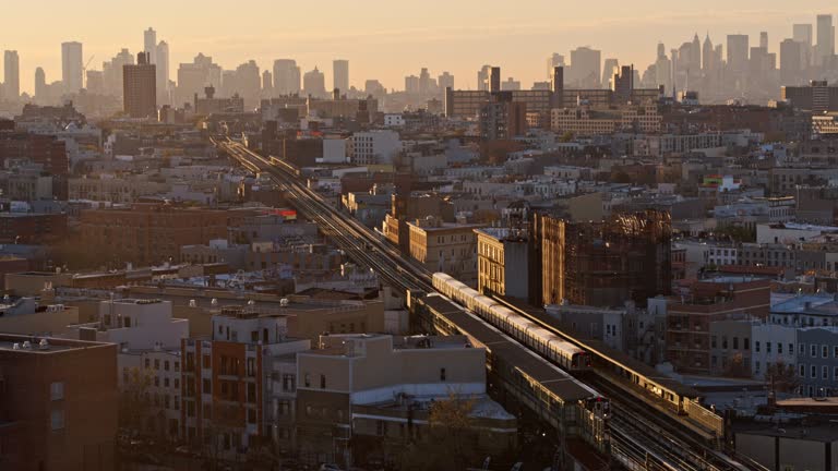 Distant view on Downtown Brooklyn and Manhattan over the residential district. A train is riding on an elevated subway line along Myrtle avenue toward Downtown Brooklyn in the evening. Drone video with the forward-panning camera motion.