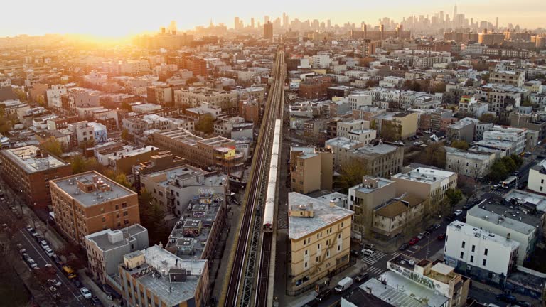 A train is riding across the residential district in Brooklyn, New York, on the sunset. Drone video with the forward and tilting-up accelerated camera motion.