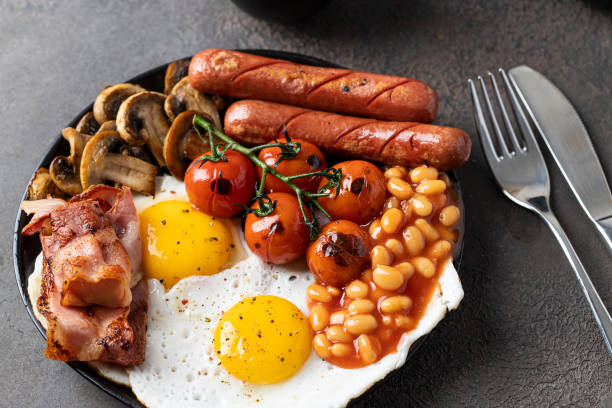 istock Typical English breakfast on dark concrete background. Fried eggs, sausages, bacon, beans, mushrooms, toasts and butter, coffee and orange juice. Copy space 1303768446