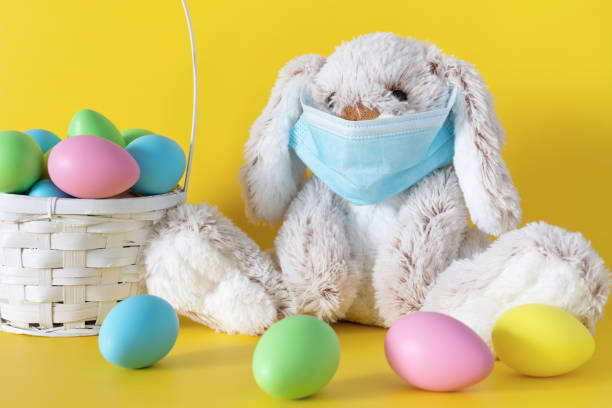 istock Easter bunny in mask and colored eggs in basket on yellow background. Ester holiday during corona virus concept. 1303791663