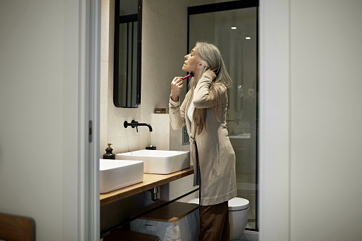 View through bathroom doorway of Caucasian woman with long gray hair wearing casual clothing and applying blush to her face.