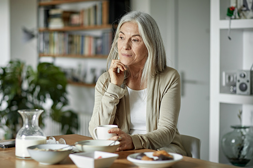Partial front view of Caucasian woman with long gray hair wearing casual clothing and sitting at dining table in modern apartment looking away from camera.