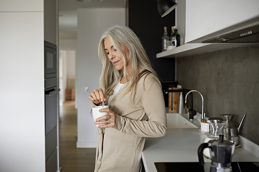 Partial side view of relaxed woman with long gray hair wearing casual clothing leaning against kitchen counter and preparing coffee in mug.