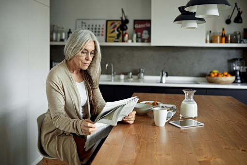 Retired Caucasian woman with long gray hair wearing casual clothing and enjoying a leisurely breakfast with coffee at dining room table in modern apartment.