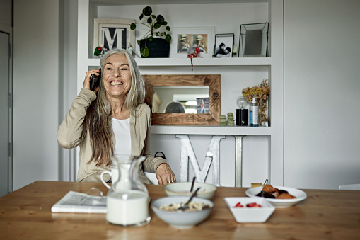 Partial front view of relaxed Caucasian woman wearing casual clothing and sitting at dining table enjoying phone call with friend in modern apartment.