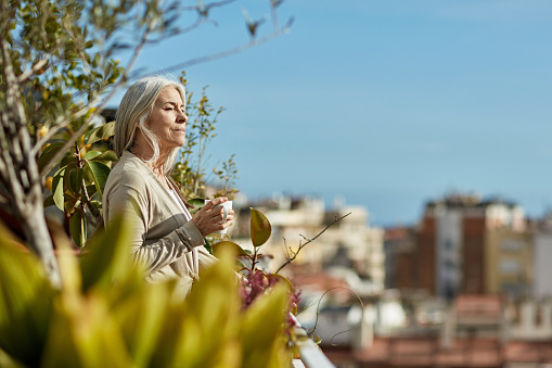 Partial side view of Caucasian woman with long gray hair wearing casual clothing and standing with coffee on apartment deck admiring view of Barcelona.
