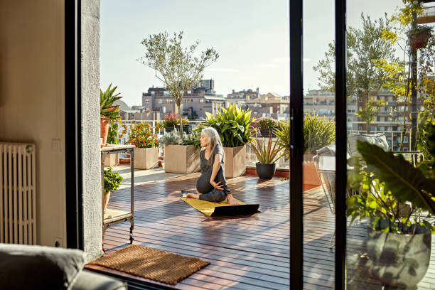 Retired Senior Woman Enjoying Yoga on Sunny Outdoor Deck Full length front view of Caucasian woman with long gray hair photographed through open sliding glass door on exercise mat in seated spinal twist position. elder plant stock pictures, royalty-free photos & images