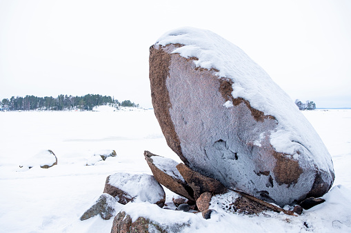 Huge granite stone. The coastline of the bay of the sea bay covered with ice. Winter nature landscape of northern Europe. Pine forest and granite boulder on the shore.