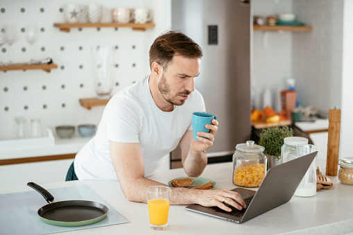 Young man eating breakfast and reading the news online. stock photo. Shadow DOF. Developed from RAW; retouched with special care and attention; Small amount of grain added for best final impression. 16 bit Adobe RGB color profile.