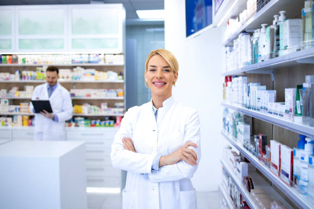 薬局店やドラッグストアに腕を組んで立っている笑顔の女性ブロンド薬剤師の肖像画。薬と背景の棚で。 - pharmacy pharmacist smiling pill ストックフォトと画像
