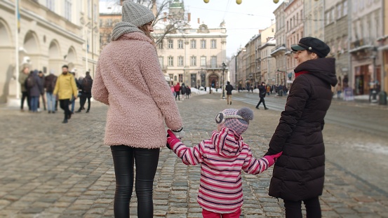 Lesbian couple tourists holding hands with adoption child girl kid stay on city center street during winter holidays vacation. Rear view of LGBT woman family with kid walking, talking outdoors