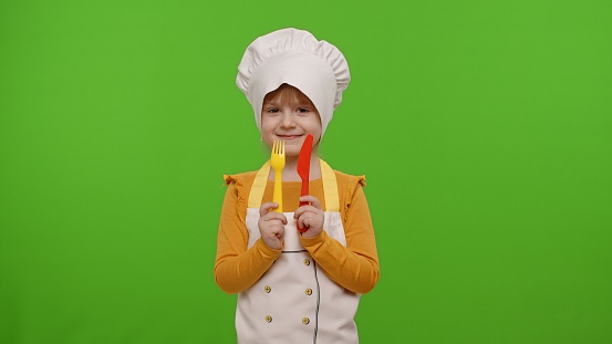 Funny Caucasian preschooler child girl dressed cook chef baker in apron and hat with plastic fork and knife fooling around, making faces on chroma key background. Cooking school, children education