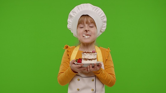 Little child girl kid dressed as professional cook chef eating tasty handmade strawberry cake isolated on chroma key background. Nutrition, cooking food school, education. Fun and humor