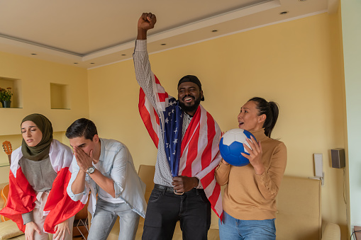 Four Multi-Ethnic Young Friends are Spending a Nice Time Together in Their Comfortable Apartment, Watching a Soccer Game, and Cheering Using an American and Canadian Flag.