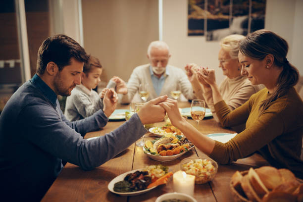 Multi-generation family holding hands and praying during lunch in dining room. Grateful extended family saying grace while sitting at dining table and holding hands. saying grace stock pictures, royalty-free photos & images