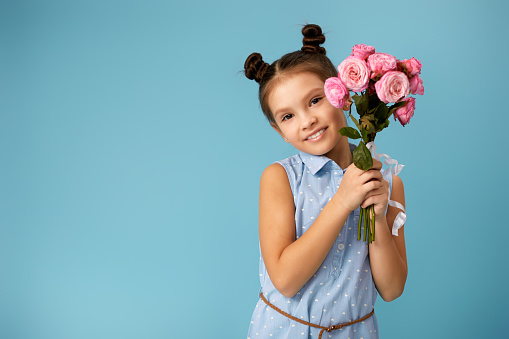 adorable smiling little girl holding bouquet of pink roses on blue background. Copy space for text.